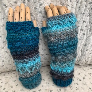 May include: Handmade fingerless gloves in blue, teal, and grey. The crochet gloves have a textured design with a ribbed cuff. They are displayed on wooden hand forms. The yarn has a subtle colour gradient. The gloves are approximately 20 cm long.