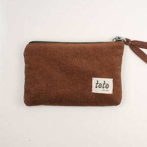 May include: A brown corduroy pouch with a black zip and a fabric pull. The pouch features a small rectangular label with the text "toto the label" in a handwritten style font. The pouch is rectangular and has a ribbed texture.