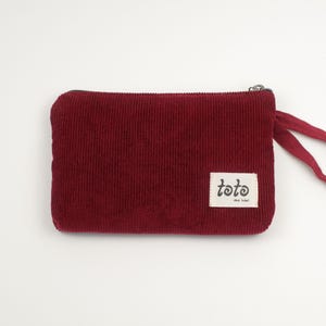 May include: A burgundy corduroy pouch with a zipper closure and a small fabric tag that reads "tote the label". The pouch has a rectangular shape with rounded corners and a matching burgundy fabric pull.