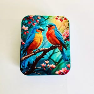Może przedstawiać: Kwadratowe pudełko z blachy z kolorowym wzorem przedstawiającym dwa niebieskie ptaki z pomarańczowymi i czerwonymi akcentami, które siedzą na gałęzi z różowymi kwiatami. Tło to niebieski i zielony gradient z białymi gwiazdami.