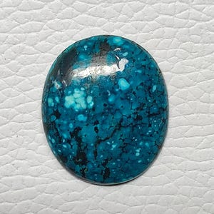 Pode incluir: Uma pedra preciosa turquesa de formato oval com uma cor azul-esverdeada vibrante e um intrincado padrão de matriz preta em toda a superfície. A pedra tem um acabamento liso e polido com um brilho lustroso que reflete a luz na parte superior.
