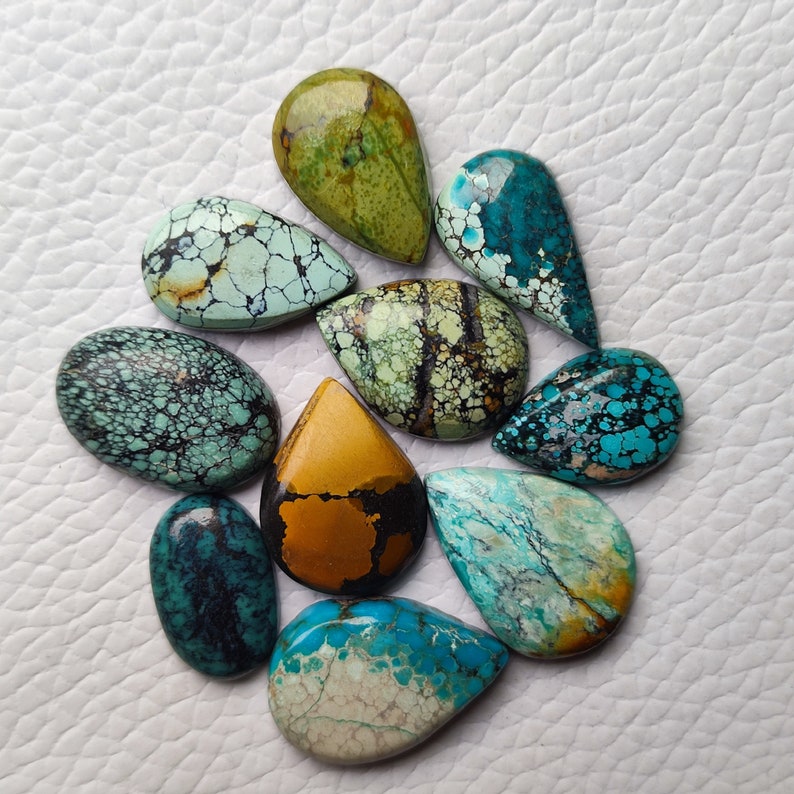 Op de afbeelding: Een verzameling van tien traanvormige turquoise cabochons in verschillende tinten blauw, groen en bruin. De stenen hebben een gevlekte, gemarmerde uitstraling met een mix van zwarte en witte aderen.