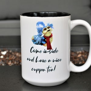 May include: White ceramic mug with a black rim. The mug features a blue and white illustration of a furry creature with a red scarf. The text on the mug reads "Come inside and have a nice cuppa tea!"