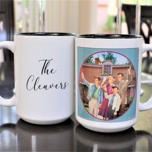 May include: Two white ceramic mugs with black rims. One mug features the text "The Cleavers" in black script. The other mug has a colorful image of a family standing in front of a house.