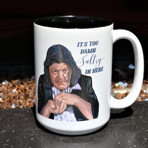 May include: White ceramic coffee mug with a black rim. The mug features a cartoon illustration of a woman with long dark hair and a serious expression. The text "It's too damn sultry in here" is printed in blue on the mug.