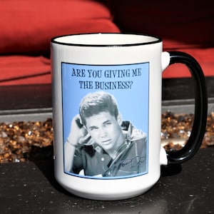 May include: White ceramic mug with a black rim and handle. The mug features a black and white image of a man with the text "Are you giving me the business?" printed on the front.
