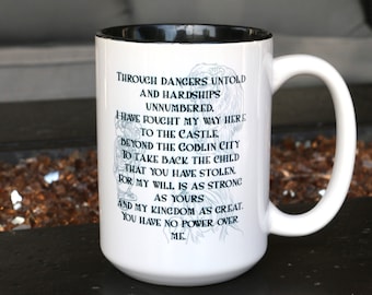 Puede incluir: Taza de cerámica blanca con borde negro. La taza presenta una ilustración en blanco y negro de un guerrero con una cita que dice: "Through dangers untold and hardships unnumbered. I have fought my way here to the castle. Beyond the goblin city to take back the child that you have stolen. For my will is as strong as yours and my kingdom as great. You have no power over me."