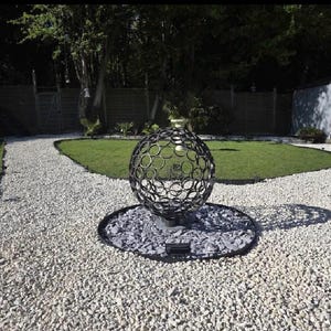 Puede incluir: Una escultura esférica decorativa de metal negro con un diseño circular, colocada sobre un lecho de piedras grises. La escultura está en un jardín con un camino de grava y hierba verde.