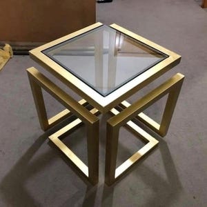 May include: A square side table featuring a gold-coloured metal frame with a geometric, open-structure design and a clear glass top. The frame consists of four separate, angular leg sections that support the square glass surface from beneath.