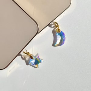 May include: Two gold-colored smartphones with black camera lenses are displayed. Each phone has a small, iridescent charm attached, one a star and the other a crescent moon. The charms are suspended by gold-colored clasps.
