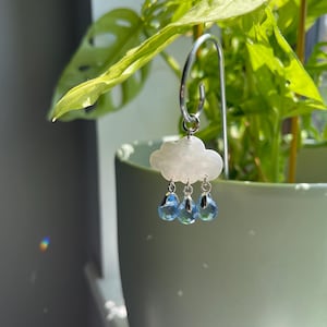 Puede incluir: Un colgante de pendiente en forma de nube con tres colgantes de cristal azul en forma de l&aacute;grima. El colgante est&aacute; suspendido de un aro plateado. El fondo presenta una planta verde en una maceta verde claro.