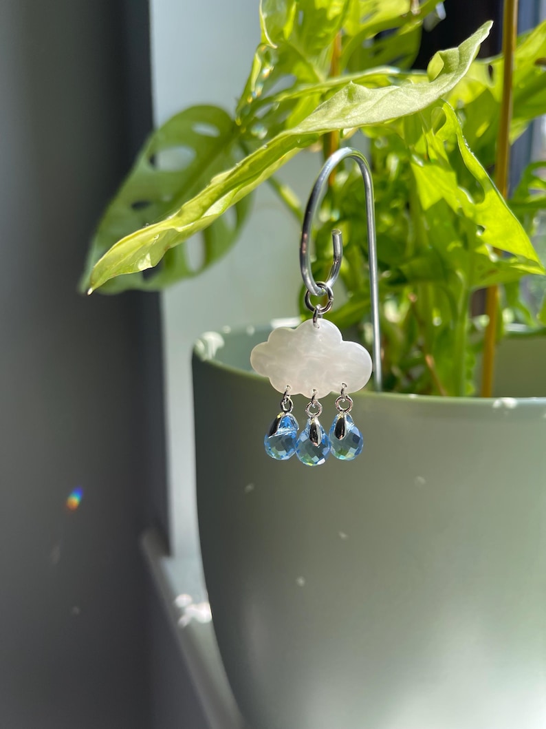 Puede incluir: Un colgante de pendiente en forma de nube con tres colgantes de cristal azul en forma de l&aacute;grima. El colgante est&aacute; suspendido de un aro plateado. El fondo presenta una planta verde en una maceta verde claro.