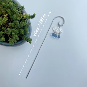 Puede incluir: Un marcap&aacute;ginas plateado con un dise&ntilde;o de nube y gotas de lluvia. La nube es blanca y texturizada, con tres cuentas azules en forma de gota de lluvia. El marcap&aacute;ginas mide 20 cm de largo.