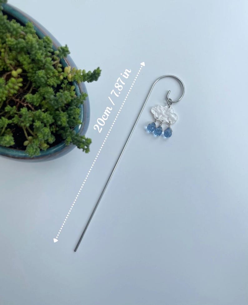 Puede incluir: Un marcap&aacute;ginas plateado con un dise&ntilde;o de nube y gotas de lluvia. La nube es blanca y texturizada, con tres cuentas azules en forma de gota de lluvia. El marcap&aacute;ginas mide 20 cm de largo.