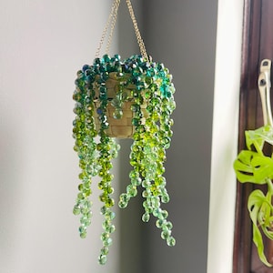 Op de afbeelding: Een hangende plantenpot, versierd met groene en blauwe kralen, hangend aan een gouden ketting. De pot is gemaakt van een licht geweven materiaal. Strengen groene kralen hangen naar beneden, wat een waterval-effect creëert.