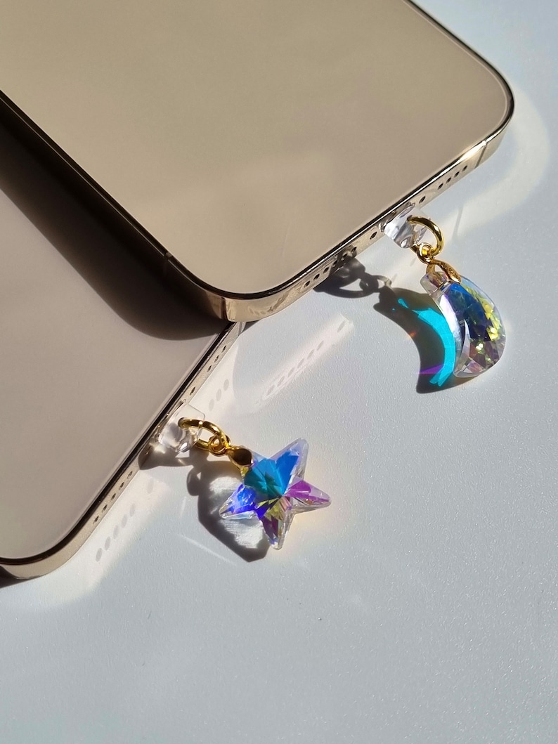 May include: Two gold-colored smartphones with iridescent crystal charms. One charm is a star, the other a crescent moon. Both charms have gold-colored metal accents and are attached to the phone's charging port.