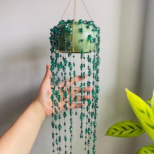 May include: A hanging planter featuring a light green ceramic bowl suspended by gold-toned chains. Numerous strands of dark green faceted beads drape downward from the rim, creating a cascading effect that mimics a trailing plant's foliage.