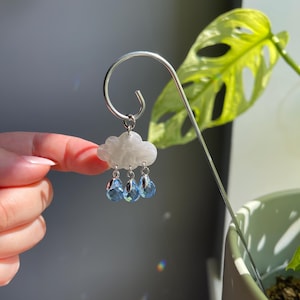 Puede incluir: Una estaca plateada para plantas con un dise&ntilde;o de nube y gotas de lluvia. La nube es blanca y las gotas de lluvia son cuentas azules con forma de cristal. La estaca est&aacute; curvada en la parte superior.
