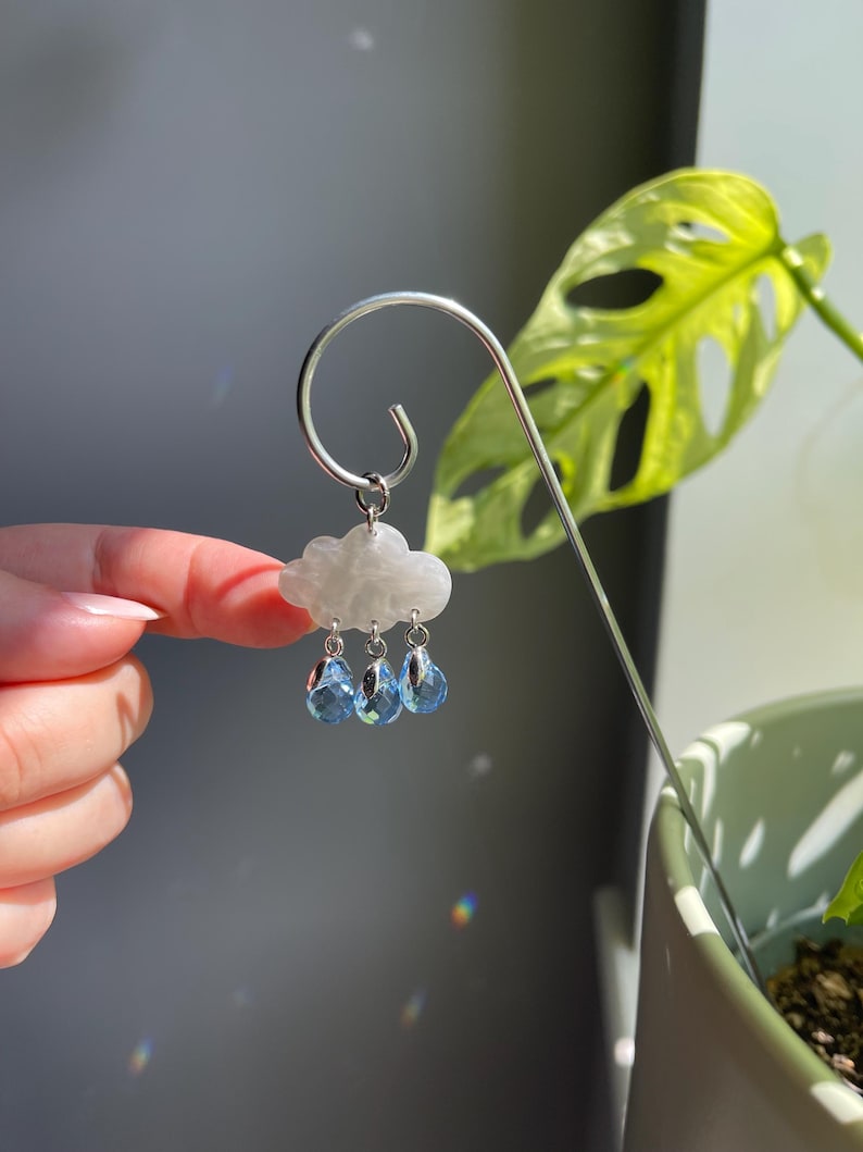 Puede incluir: Una estaca plateada para plantas con un dise&ntilde;o de nube y gotas de lluvia. La nube es blanca y las gotas de lluvia son cuentas azules con forma de cristal. La estaca est&aacute; curvada en la parte superior.
