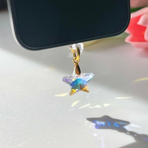 May include: A clear, star-shaped phone charm with iridescent facets hangs from a gold-colored loop. The charm is suspended near a black phone, casting a star-shaped shadow on a white surface. The charm's design catches and reflects light.