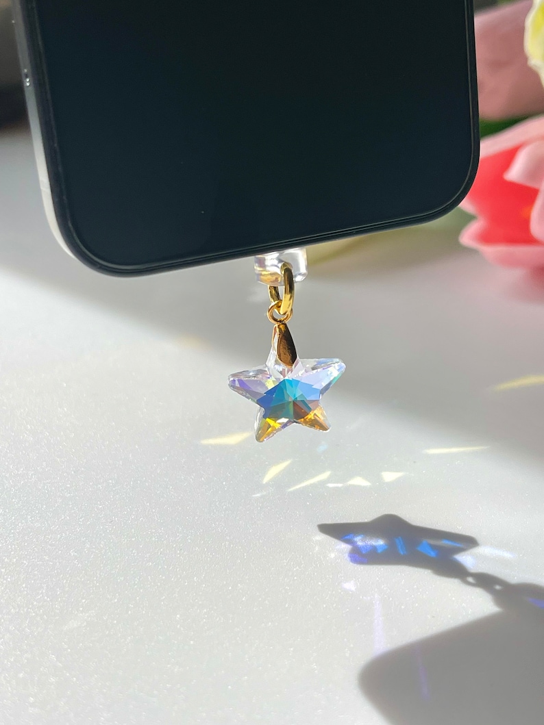 May include: A clear, star-shaped phone charm with iridescent facets hangs from a gold-colored loop. The charm is suspended near a black phone, casting a star-shaped shadow on a white surface. The charm's design catches and reflects light.