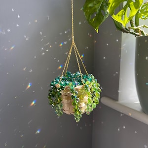 May include: A hanging planter with a woven basket and a cascade of green and clear beads. The planter is suspended by a gold chain. The background is a gray wall with light reflections and a potted plant.