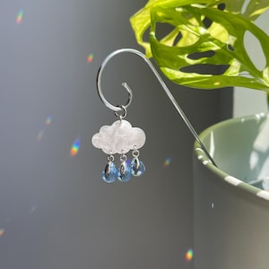 Puede incluir: Marcap&aacute;ginas plateado con un charm de nube blanca y tres gotas de lluvia de cristal azul en forma de l&aacute;grima. La nube tiene una superficie texturizada. El marcap&aacute;ginas cuelga frente a una planta verde.