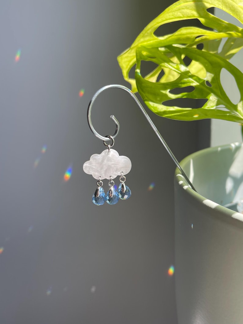 Puede incluir: Marcap&aacute;ginas plateado con un charm de nube blanca y tres gotas de lluvia de cristal azul en forma de l&aacute;grima. La nube tiene una superficie texturizada. El marcap&aacute;ginas cuelga frente a una planta verde.