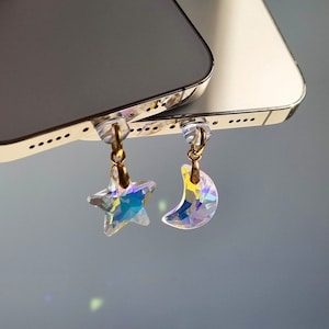 May include: Iridescent star and crescent moon charms with gold-colored accents. The charms are attached to clear plastic pieces. The background is a blurred gray.