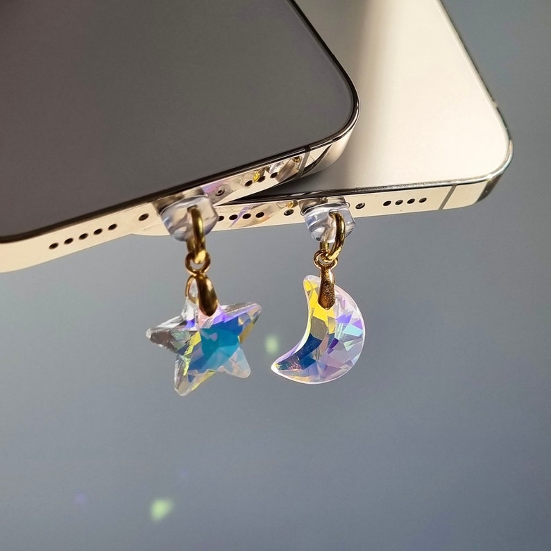 May include: Iridescent star and crescent moon charms with gold-colored accents. The charms are attached to clear plastic pieces. The background is a blurred gray.