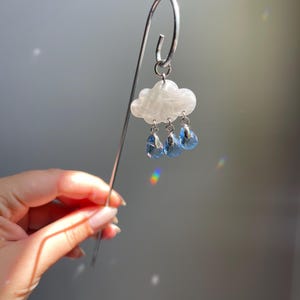 Puede incluir: Marcap&aacute;ginas plateado con un charm de nube blanca y tres colgantes de gota de lluvia azules. La nube tiene un aspecto veteado. El marcap&aacute;ginas se sostiene frente a un fondo gris.