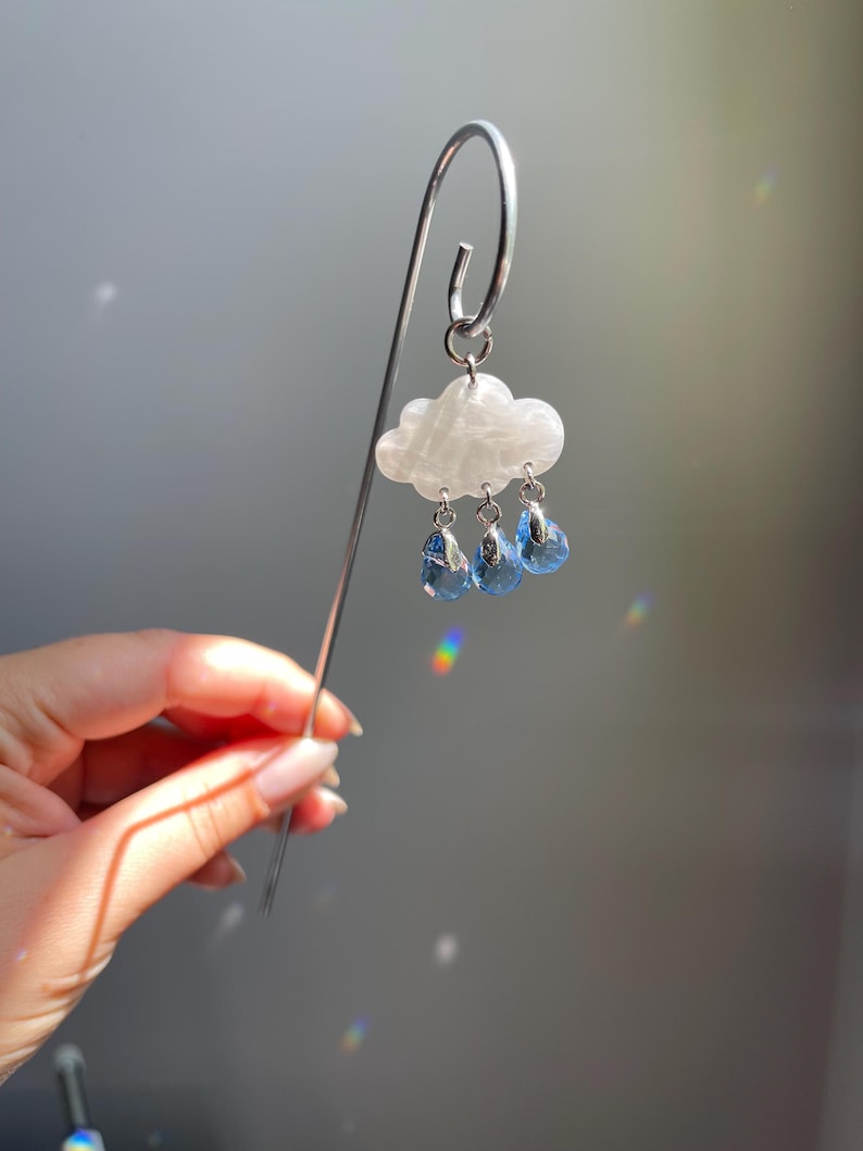 Puede incluir: Marcap&aacute;ginas plateado con un charm de nube blanca y tres colgantes de gota de lluvia azules. La nube tiene un aspecto veteado. El marcap&aacute;ginas se sostiene frente a un fondo gris.