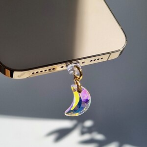 May include: A gold-colored phone with a crystal crescent moon charm hanging from the bottom. The charm is iridescent, reflecting multiple colors. The phone is angled, showing its edge and the charm's shadow.