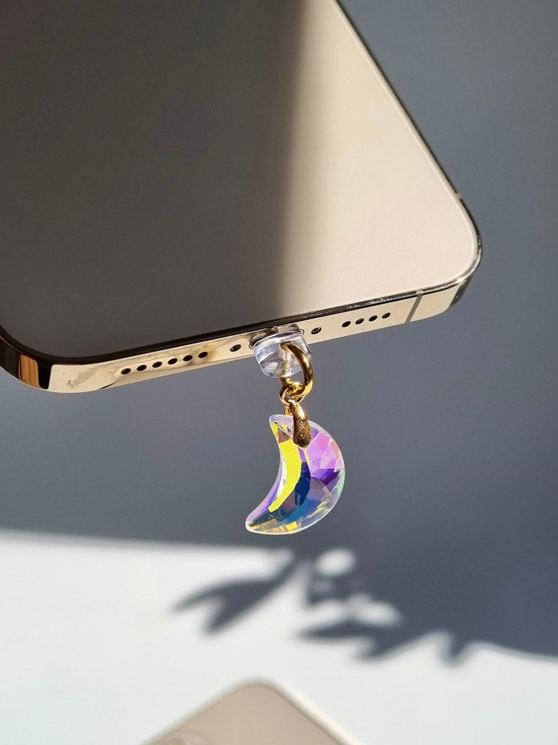 May include: A gold-colored phone with a crystal crescent moon charm hanging from the bottom. The charm is iridescent, reflecting multiple colors. The phone is angled, showing its edge and the charm's shadow.