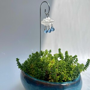 Puede incluir: Una peque&ntilde;a maceta de cer&aacute;mica turquesa llena de suculentas verdes. Una estaca met&aacute;lica decorativa sostiene un adorno de nube con tres cuentas azules en forma de l&aacute;grima, creando una escena de jard&iacute;n en miniatura. El adorno de nube es blanco con un acabado nacarado.