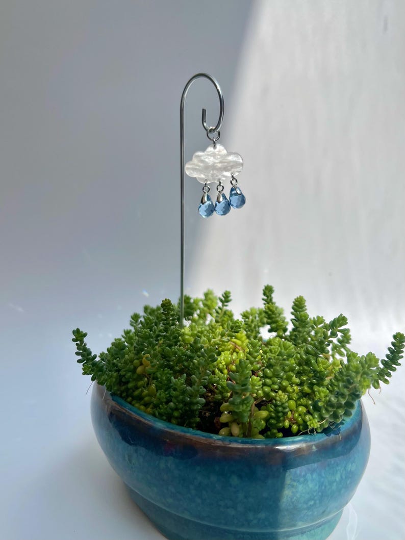 Puede incluir: Una peque&ntilde;a maceta de cer&aacute;mica turquesa llena de suculentas verdes. Una estaca met&aacute;lica decorativa sostiene un adorno de nube con tres cuentas azules en forma de l&aacute;grima, creando una escena de jard&iacute;n en miniatura. El adorno de nube es blanco con un acabado nacarado.