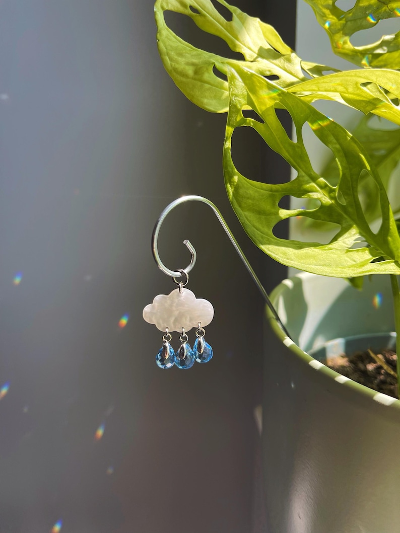 Puede incluir: Un adorno decorativo para plantas con forma de nube blanca y tres colgantes de cristal azul en forma de l&aacute;grima. El adorno est&aacute; suspendido de un gancho de metal plateado, sobre un fondo gris y una planta verde en maceta.