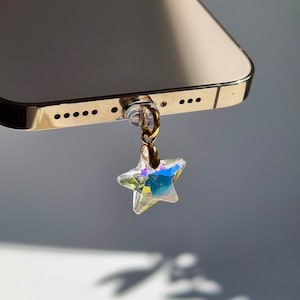 May include: A gold-colored smartphone with a clear phone charm. The charm is a star-shaped crystal with iridescent colors, attached to a gold-colored ring. The phone has a gold-colored frame.