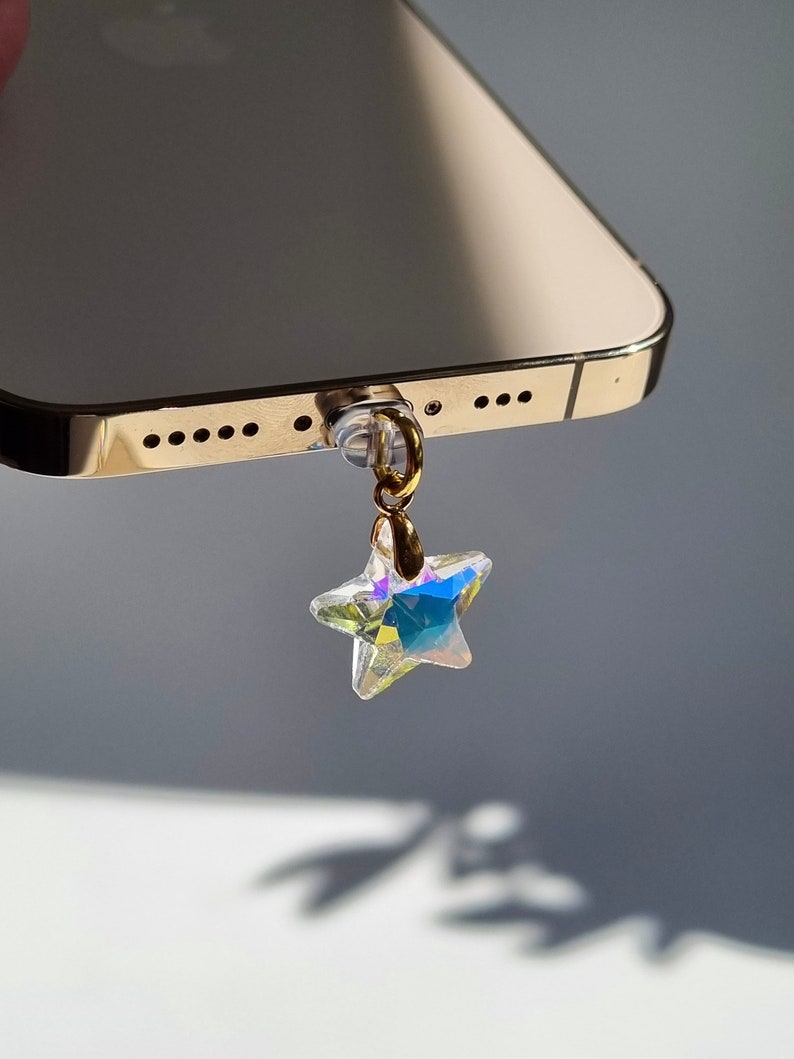 May include: A gold-colored smartphone with a clear phone charm. The charm is a star-shaped crystal with iridescent colors, attached to a gold-colored ring. The phone has a gold-colored frame.
