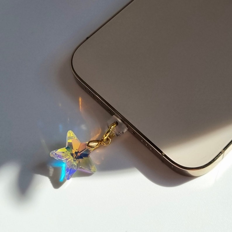 May include: A clear, iridescent star-shaped charm with a gold-colored clasp attached to a phone. The star reflects multiple colors. The phone is a light gold color.