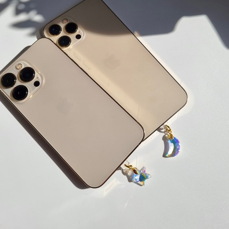 May include: Two gold-colored smartphones with black camera lenses are displayed. Each phone has a small, iridescent charm attached, one a star and the other a crescent moon. The charms are suspended by gold-colored clasps.