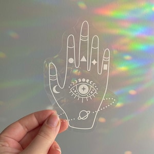 以下が含まれることがあります： 白い輪郭で描かれた目、月、星、幾何学模様のある手の形をした、透明な虹色のステッカー。ステッカーは手で持たれている。
