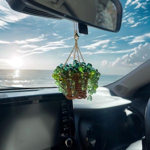 May include: A car hanging ornament featuring a small woven basket filled with green and clear beaded accents, suspended by a gold chain. The ornament hangs from the rearview mirror, with a beach and sky in the background.