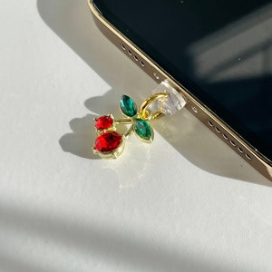 Può includere: Un ciondolo dorato con strass rossi e verdi a forma di ciliegia. Il ciondolo è attaccato a una custodia per telefono.