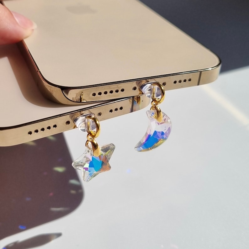 May include: A pair of iridescent crystal phone charms, one a star and the other a crescent moon, hang from the bottom of two gold-colored smartphones. The charms have gold-colored hoops and reflect multiple colors.