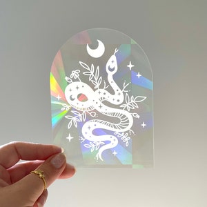 Puede incluir: Una pegatina holográfica transparente con un diseño de serpiente blanca, una luna creciente, estrellas y detalles florales. La pegatina tiene una parte superior arqueada y está sujeta por una mano.