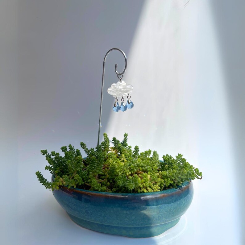 Puede incluir: Una peque&ntilde;a maceta de cer&aacute;mica turquesa llena de suculentas verdes. Un soporte de metal plateado sostiene un adorno de nube con detalles de gotas de lluvia azules, a&ntilde;adiendo un toque decorativo.