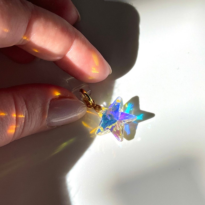 May include: A clear, iridescent star-shaped charm with a gold-colored clasp. The charm is held up against a white background, with light reflecting off the star's facets, creating a rainbow effect.