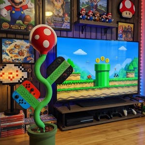 May include: A green, S-shaped plant sculpture with a red and white spotted head, rooted in a green cylindrical pot. Two game controllers are attached to the stem, one featuring blue and red buttons and the other with black and red buttons.