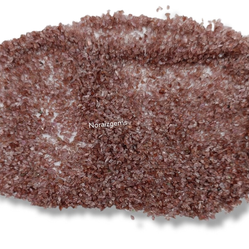 K&ouml;nnte beinhalten: Eine Nahaufnahme eines Haufens kleiner, rotbrauner Kristalle. Die Kristalle sind unregelm&auml;&szlig;ig geformt und haben eine leicht durchscheinende Optik. Die Kristalle sind auf einem wei&szlig;en Hintergrund verstreut.